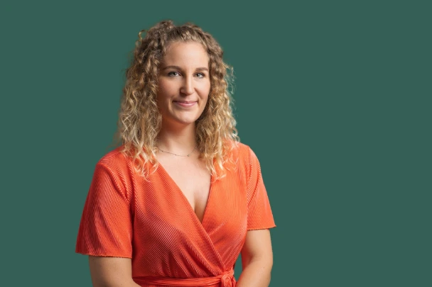 Buste de Fanny en haut rouge à manche mi courtes et fond bleu foncé, fanny se tient droite avec un sourire, les cheveux bruns à la racine et blonds au bout, cheveux mi long